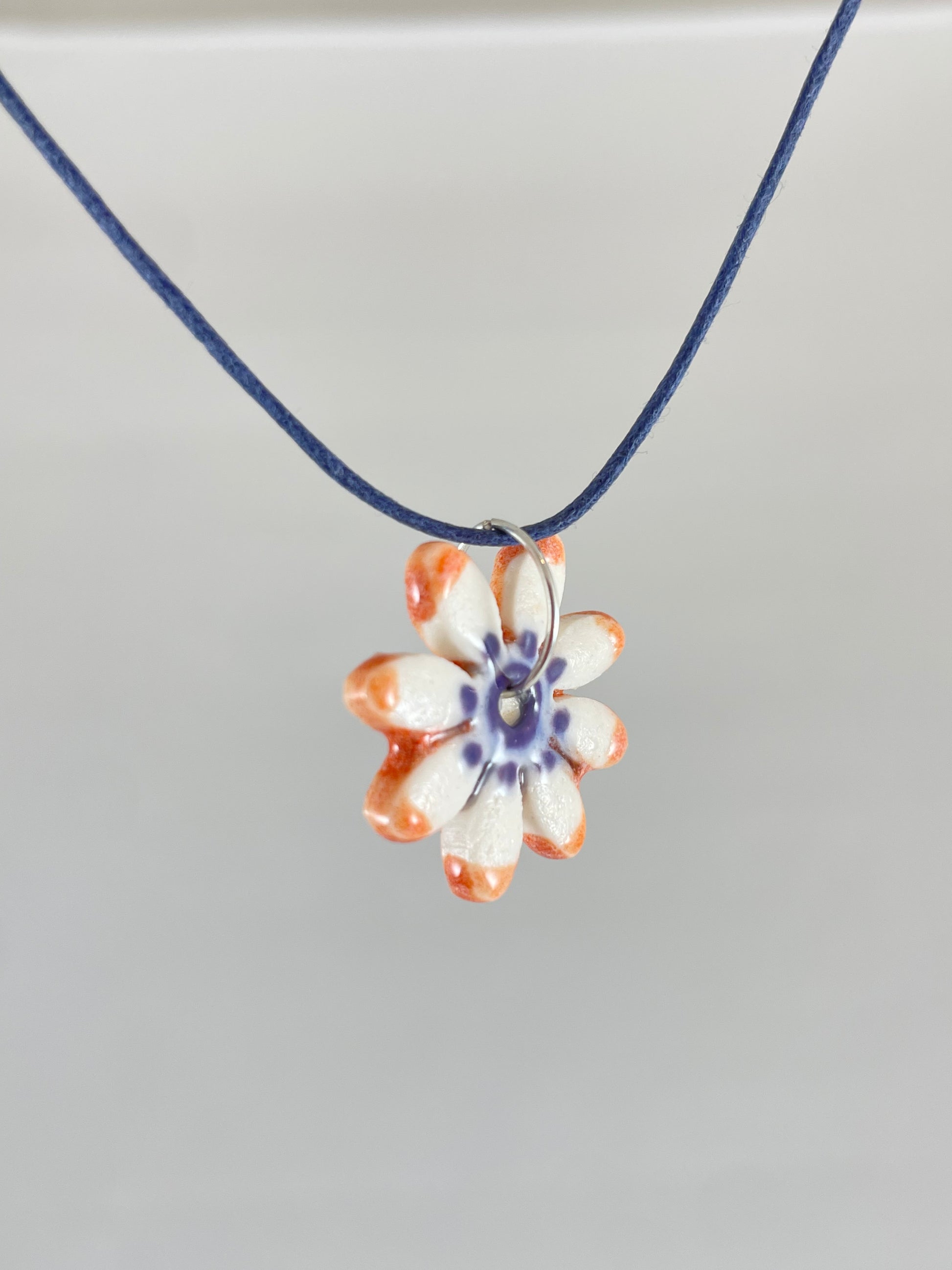 Necklace with one small daisy shaped porcelain pendant hanging on navy blue thread. Painted with lavender glaze in the centre and orange at the ends of petals. Photographed from left hand side showing thin pendant profile 