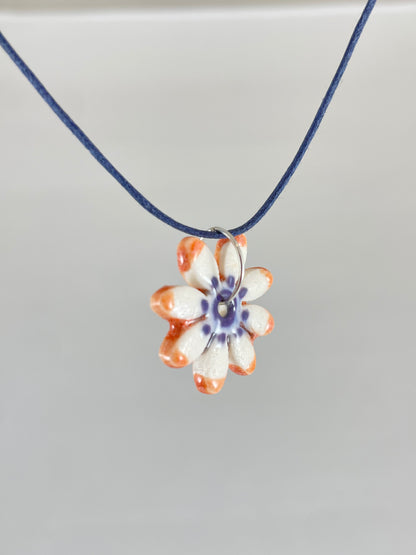 Necklace with one small daisy shaped porcelain pendant hanging on navy blue thread. Painted with lavender glaze in the centre and orange at the ends of petals. Photographed from left hand side showing thin pendant profile 