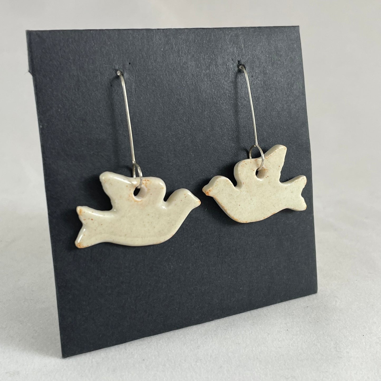 A pair of cream porcelain earrings in the shape of flying dove on a black background. Side view