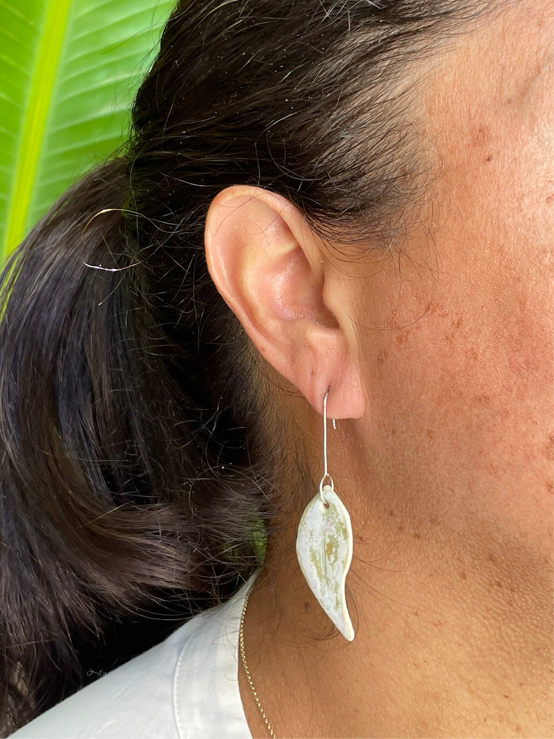 Closeup of woman wearing gum leaf earrings. Woman has olive skin and her dark brown hair is pulled back in ponytail. Sun is shining on her hair