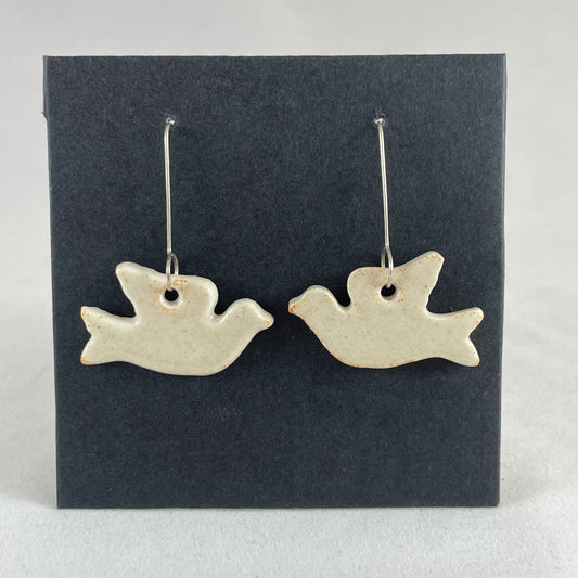 A pair of cream porcelain earrings in the shape of flying dove on a black background.