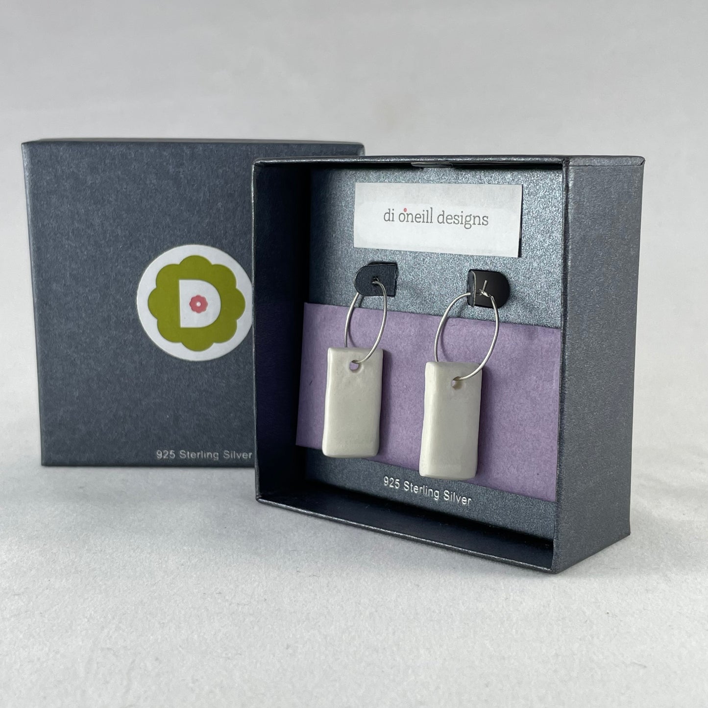 A pair of silver hoops with hand formed white porcelain pendants in a graphite coloured jewellery box. There is lilac tissue paper in the box and the box is branded with my logo and stamped with words 925 sterling silver.