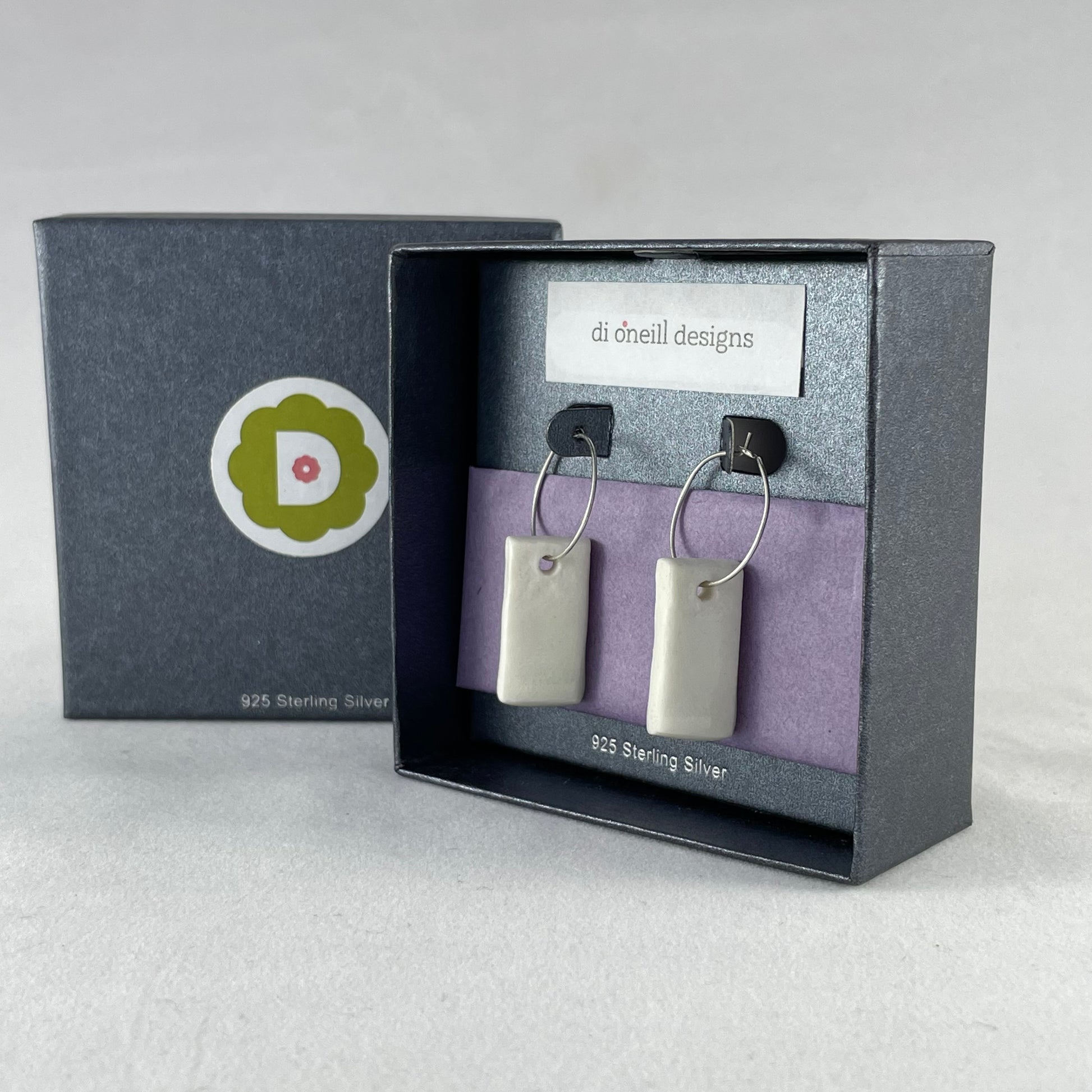 A pair of silver hoops with hand formed white porcelain pendants in a graphite coloured jewellery box. There is lilac tissue paper in the box and the box is branded with my logo and stamped with words 925 sterling silver.