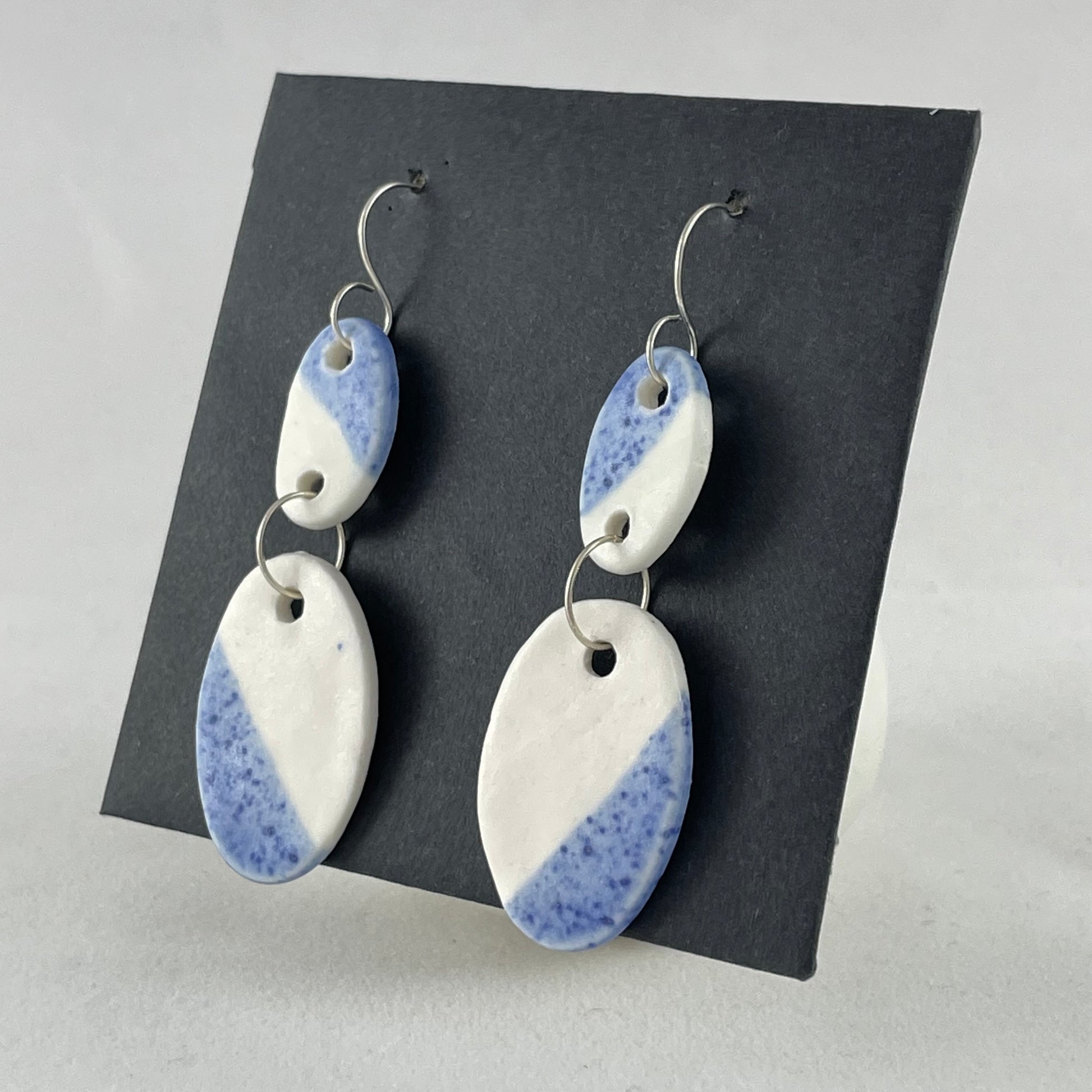 A pair of handmade porcelain earrings on a black background. Asymmetrical glaze pattern in blue and white on oval pendants. Side view