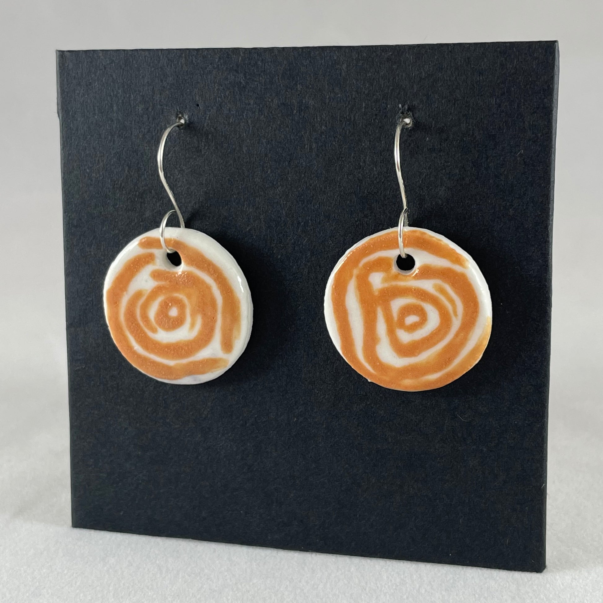 A pair of orange and white porcelain earrings hanging on black card. Loosely painted orange circles on white porcelain. Photographed from left hand side showing slim earring profile.