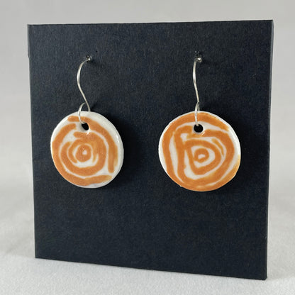 A pair of orange and white porcelain earrings hanging on black card. Loosely painted orange circles on white porcelain. Photographed from left hand side showing slim earring profile.