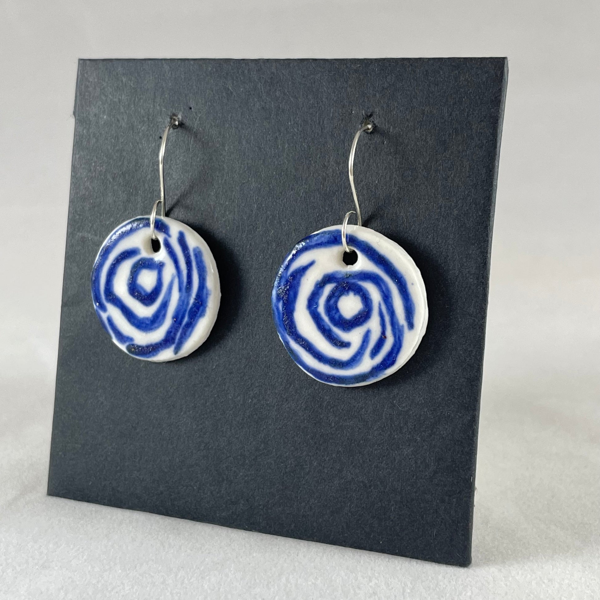 A pair of blue and white porcelain earrings hanging on black card. Simple rose pattern with loosely painted blue circles on white porcelain. Photographed from right hand side showing slim earring profile 