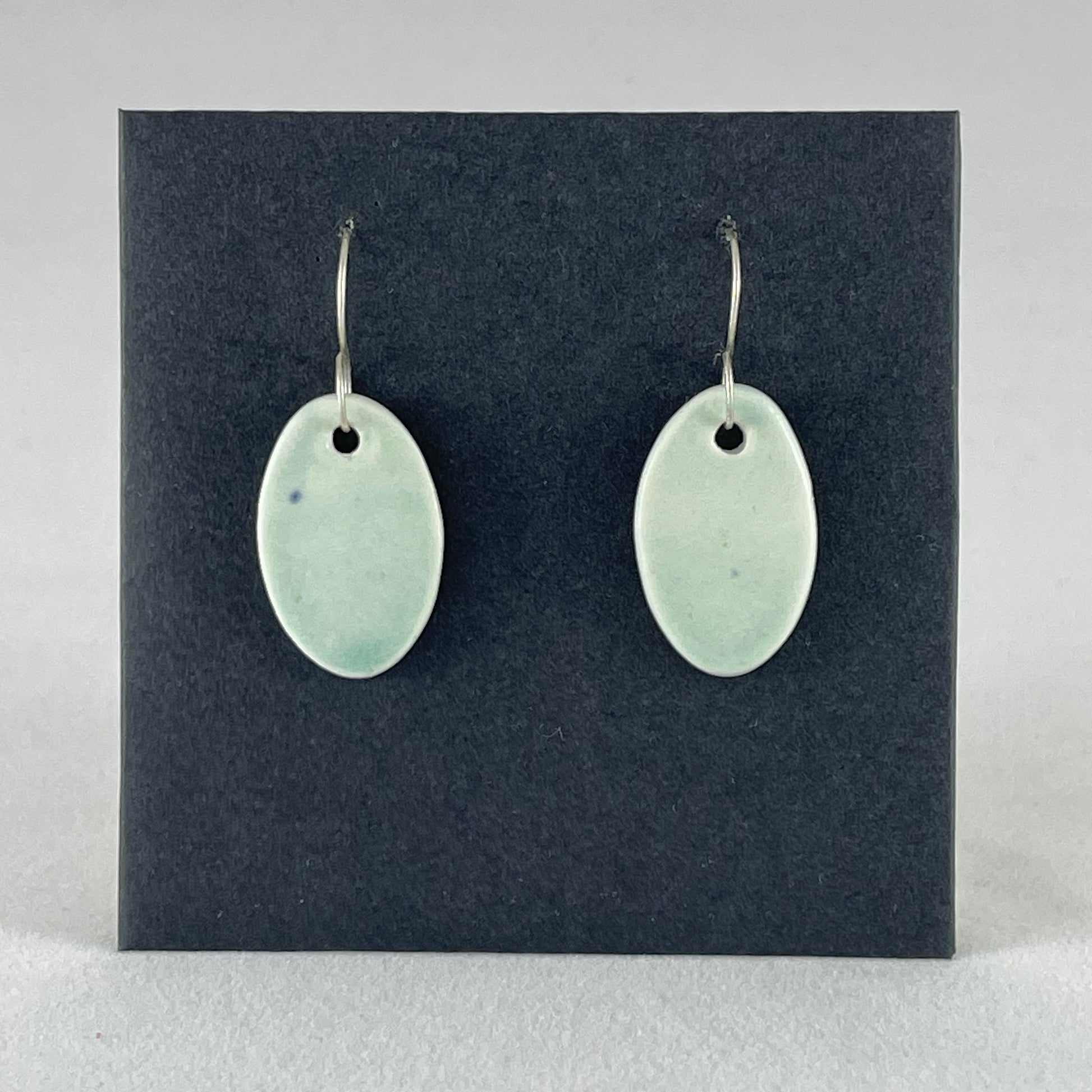Pair of very pale mint green oval shaped porcelain earrings. Pictured in front of black background. 