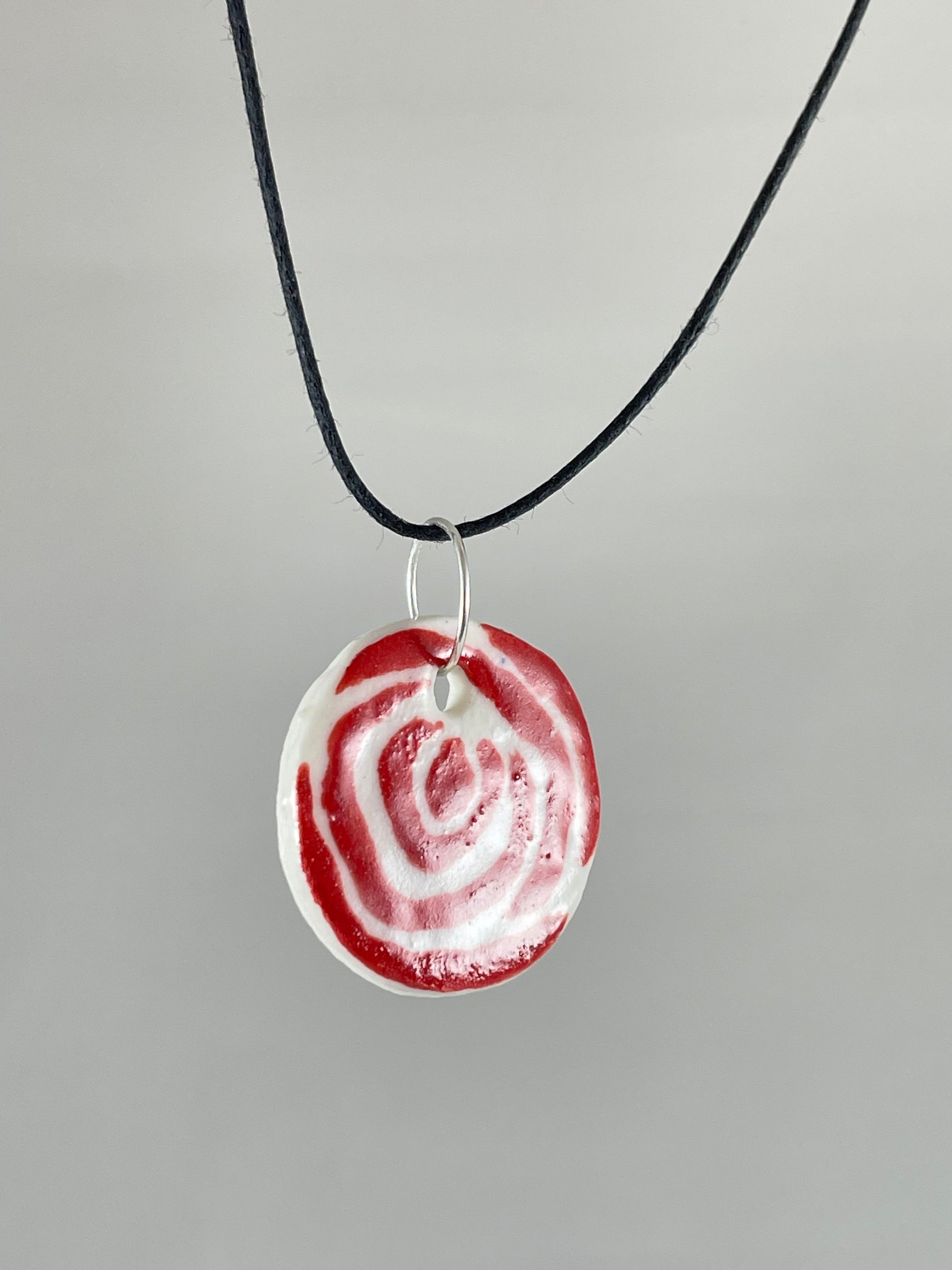 Necklace with one circular porcelain pendant on black coloured thread, hanging in front of a white background. Red rose pattern painted on white porcelain.  Photographed from left hand side to show thin profile of porcelain pendant