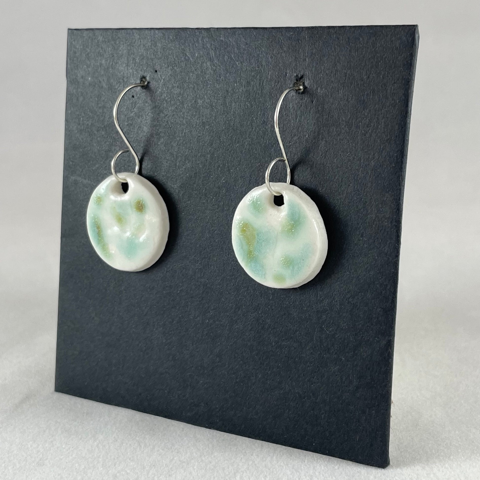 A pair of mint, olive green and white porcelain earrings hanging on black card. Photographed from right hand side showing slim earring profile.
