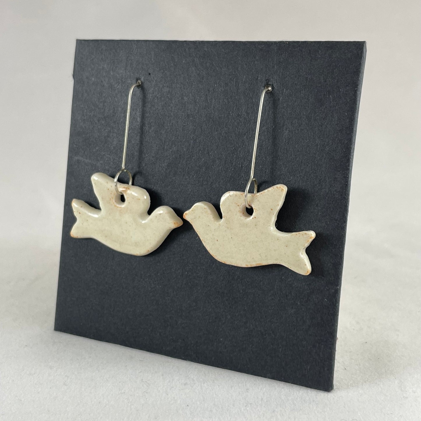 A pair of cream porcelain earrings in the shape of flying dove on a black background. Side view