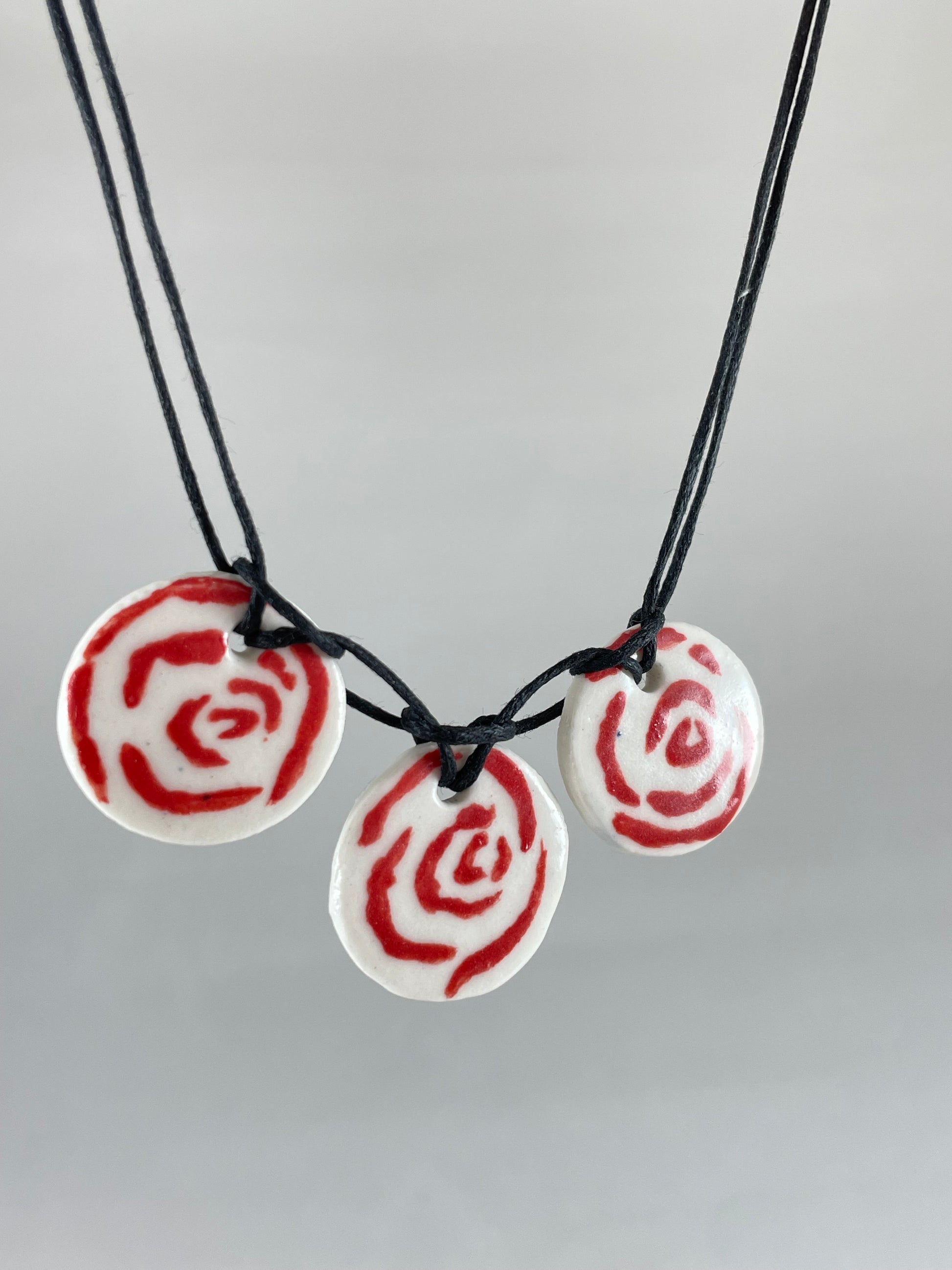 Necklace with three circular porcelain pendants on black coloured thread, hanging in front of a white background. Red rose pattern painted on white porcelain. Photographed from left side to show thin profile of porcelain discs