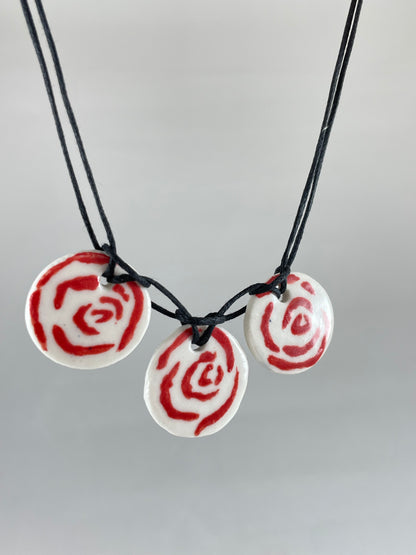 Necklace with three circular porcelain pendants on black coloured thread, hanging in front of a white background. Red rose pattern painted on white porcelain. Photographed from left side to show thin profile of porcelain discs
