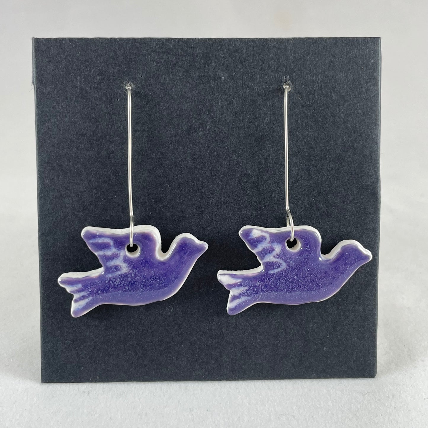 A pair of lavender porcelain earrings in the shape of flying dove on a black background.