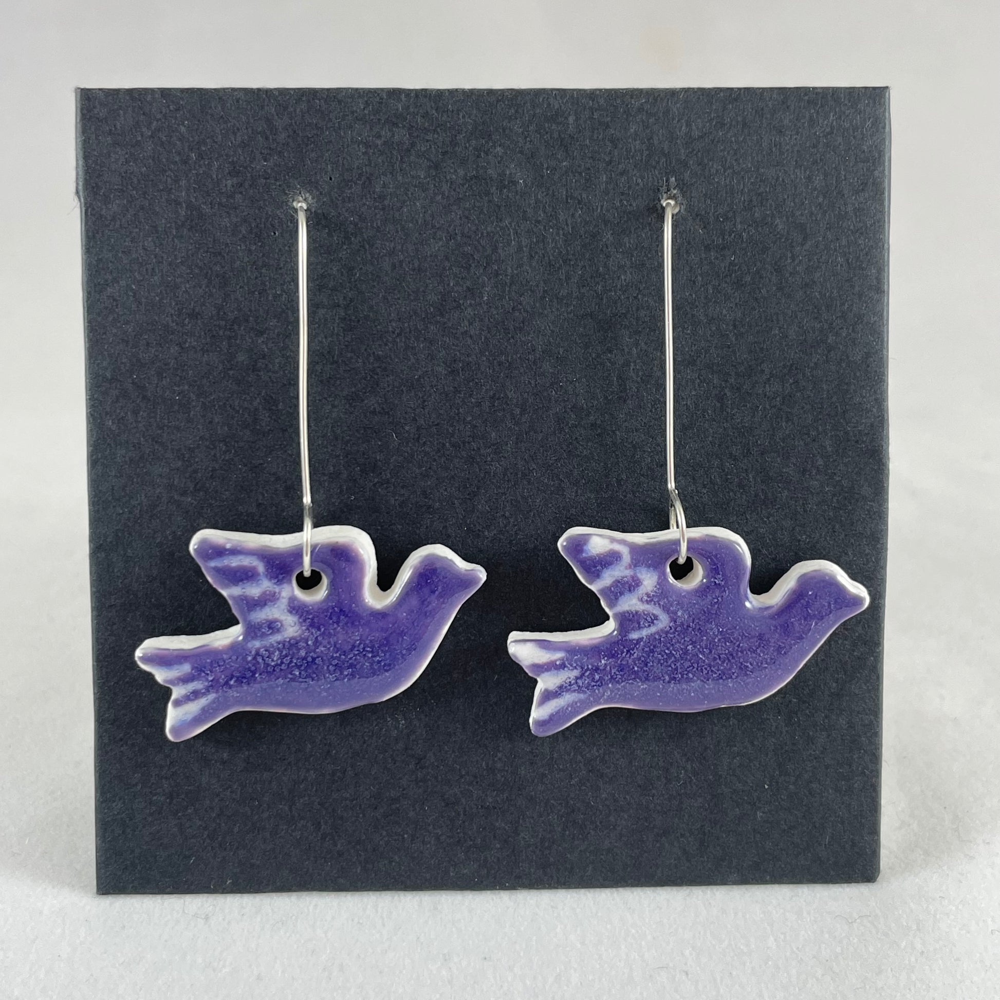 A pair of lavender porcelain earrings in the shape of flying dove on a black background.