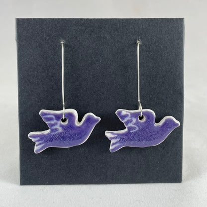 A pair of lavender porcelain earrings in the shape of flying dove on a black background.