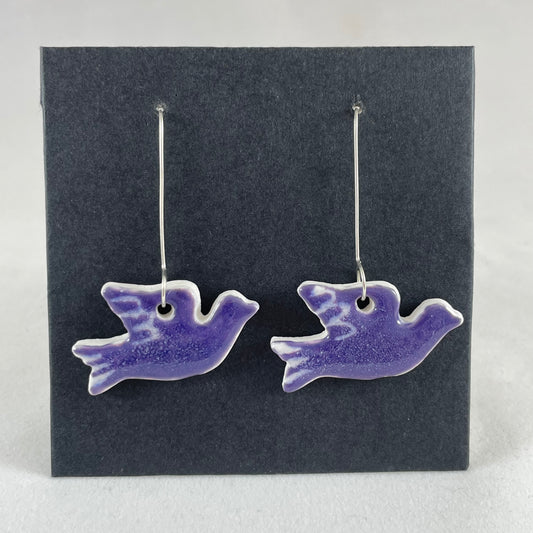 A pair of lavender porcelain earrings in the shape of flying dove on a black background.