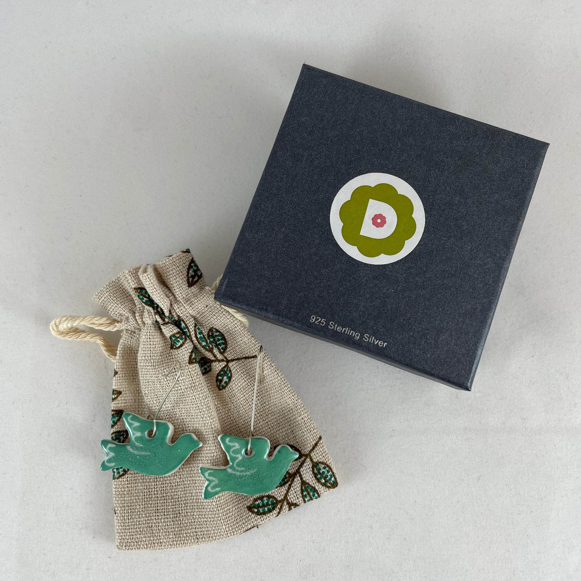 A pair of green porcelain earrings in the shape of flying dove. Photographed on a linen pouch next to jewellery box.