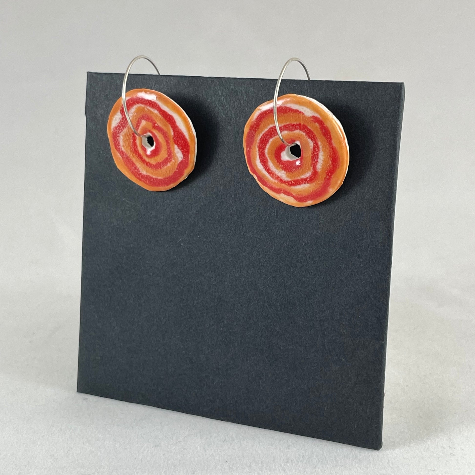 A pair of red and orange porcelain earrings on black card photographed in a white light box. Simple rose pattern. Side view from right hand side 