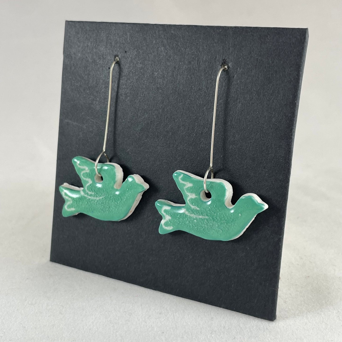 A pair of green porcelain earrings in the shape of flying dove on a black background. Side view