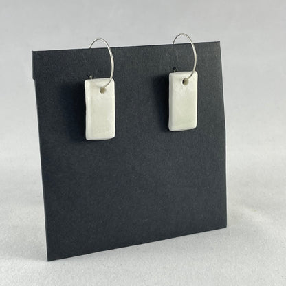 A pair of silver hoops with hand formed white porcelain pendants on a black background. Side view shot from left
