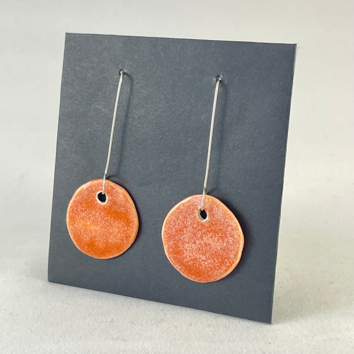 A pair of circular shaped porcelain earrings in bright orange. On sterling silver earrings hooks. Side view