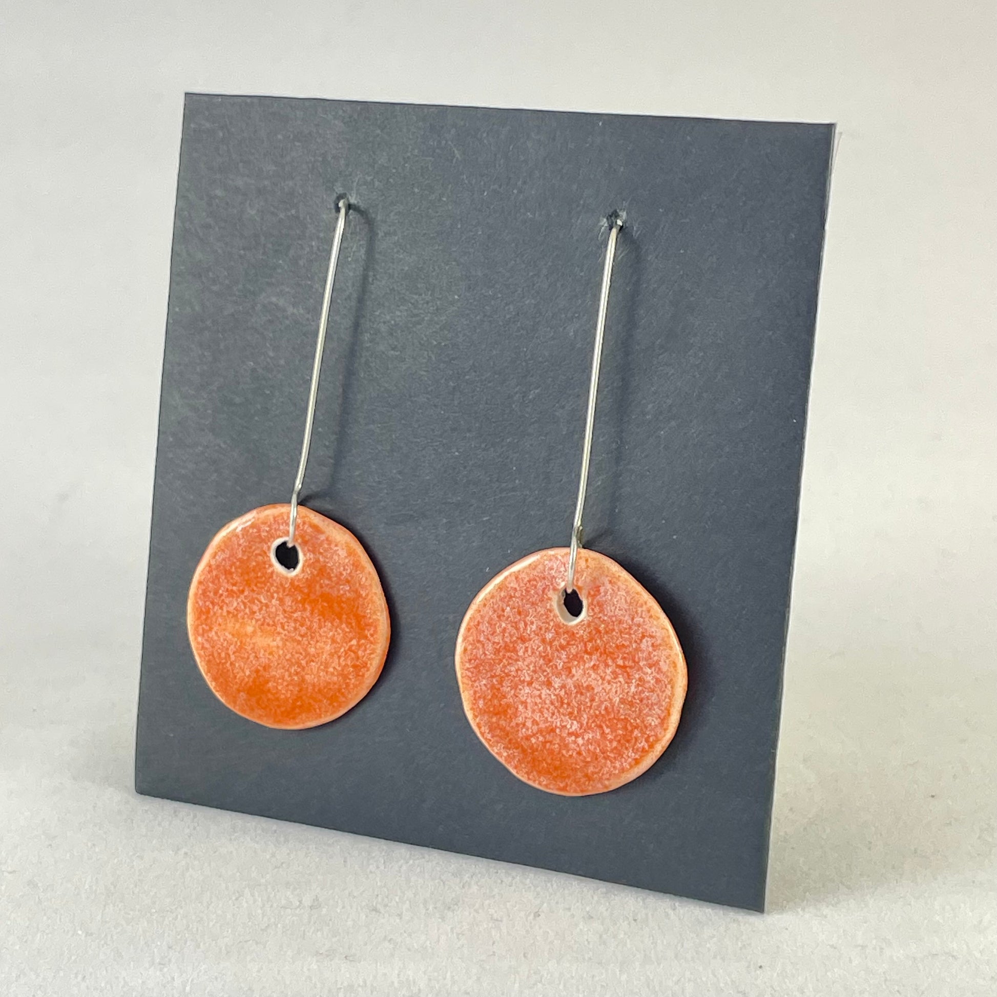 A pair of circular shaped porcelain earrings in bright orange. On sterling silver earrings hooks. Side view