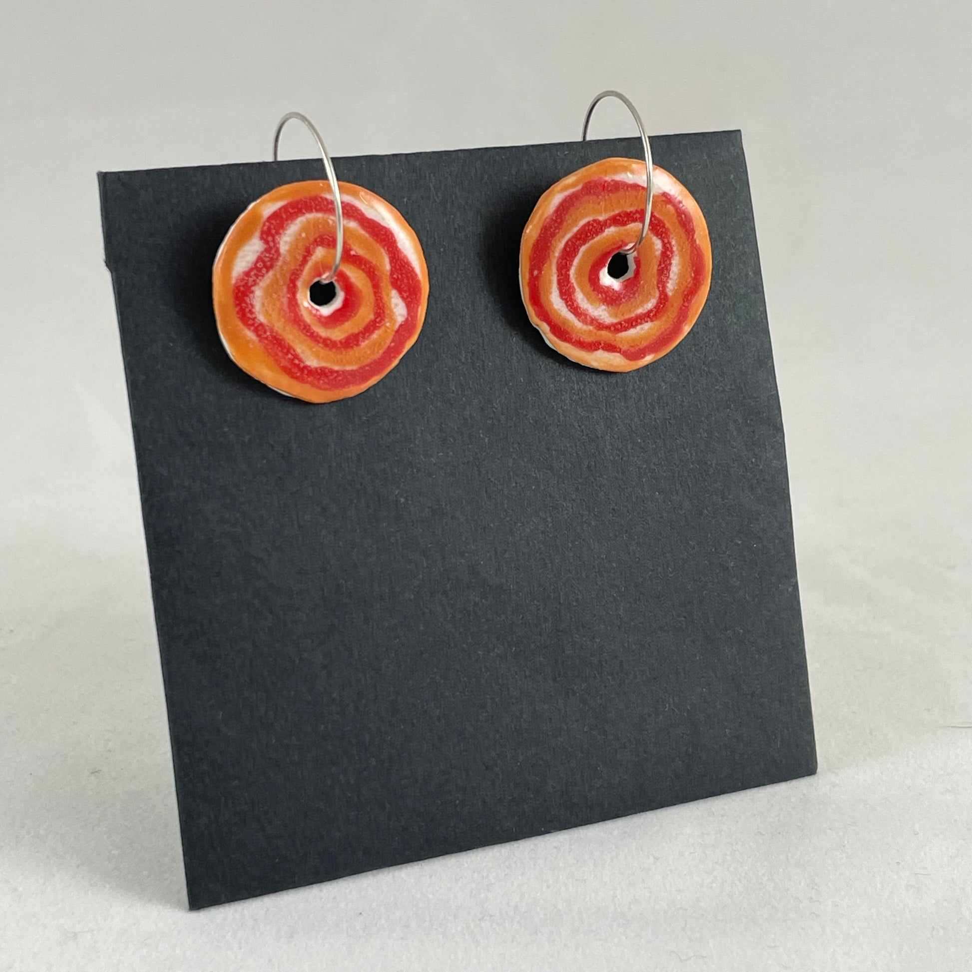 A pair of red and orange porcelain earrings on black card photographed in a white light box. Simple rose pattern. Side view from left hand side 