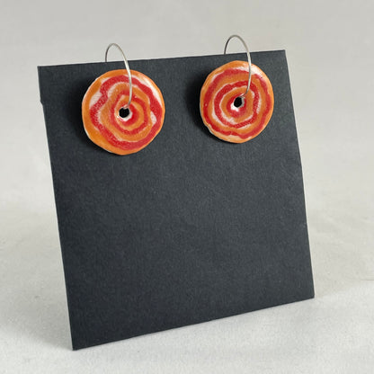 A pair of red and orange porcelain earrings on black card photographed in a white light box. Simple rose pattern. Side view from left hand side 