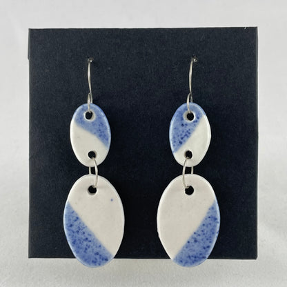 A pair of handmade porcelain earrings on a black background. Asymmetrical glaze pattern in blue and white on oval pendants.