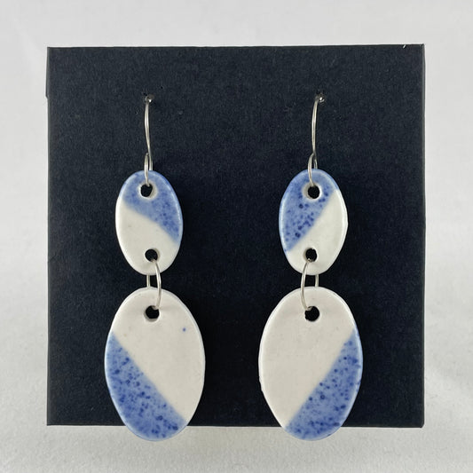 A pair of handmade porcelain earrings on a black background. Asymmetrical glaze pattern in blue and white on oval pendants.