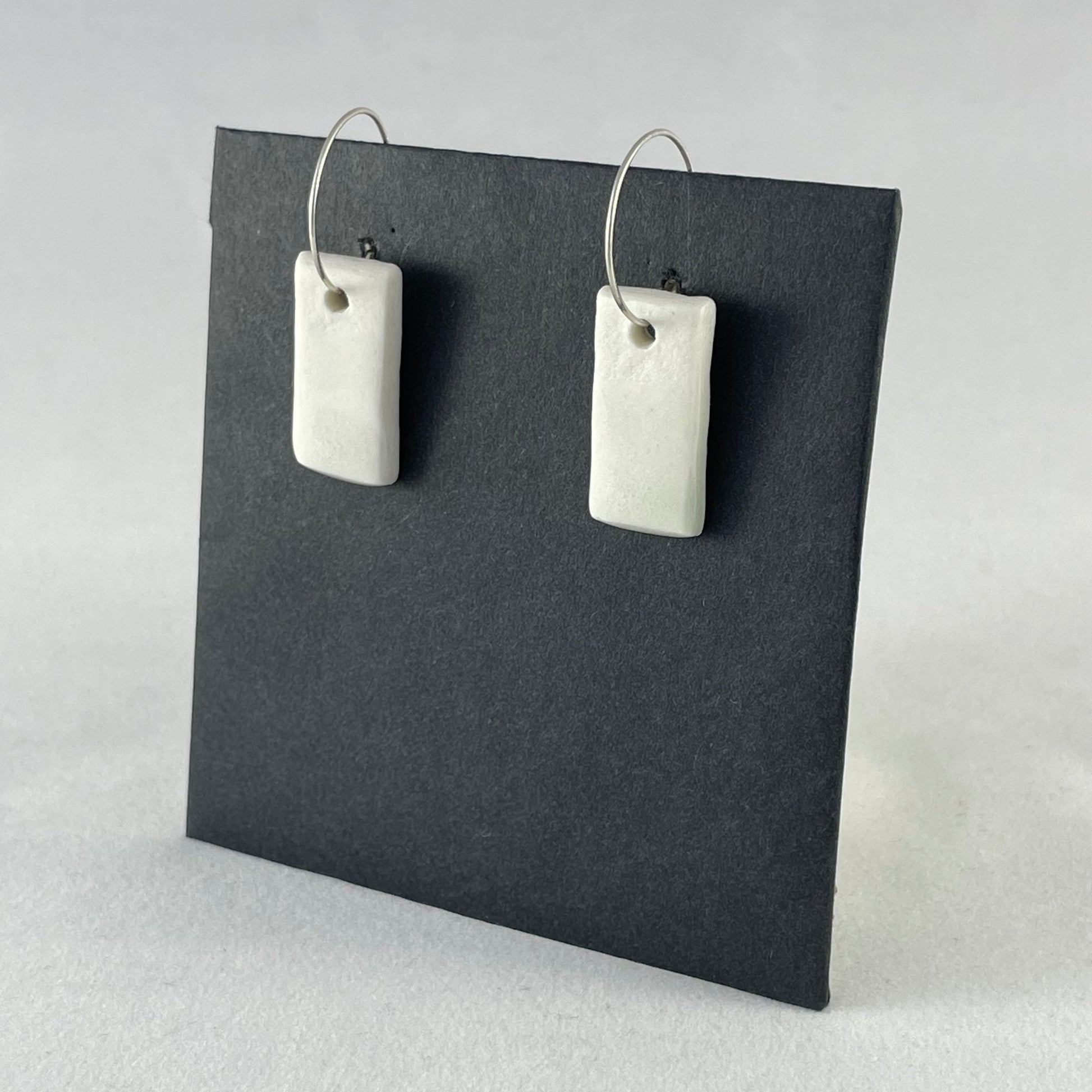 A pair of silver hoops with hand formed white porcelain pendants on a black background. Side view