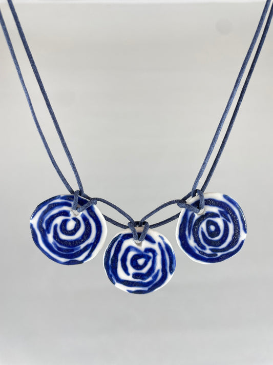 Necklace with three circular porcelain pendants on navy blue coloured thread, hanging in front of a white background. Blue rose pattern painted on white porcelain.  