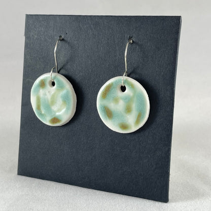 A pair of mint, olive green and white porcelain earrings hanging on black card. Photographed from right hand side showing slim earring profile.