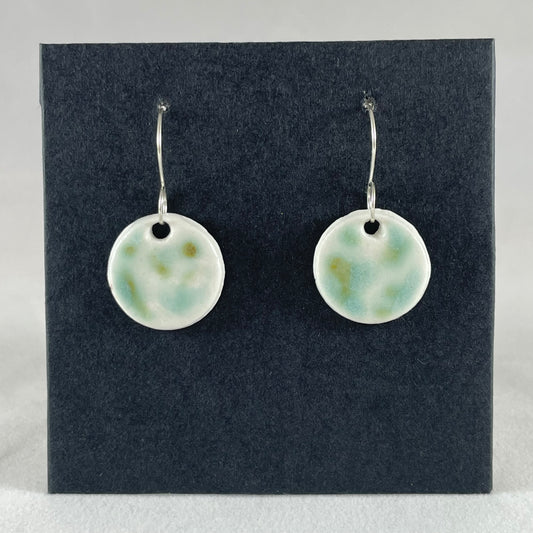 A pair of mint, olive green and white porcelain earrings hanging on black card. Splodges of glossy glaze look vaguely like a blurry camouflage pattern.