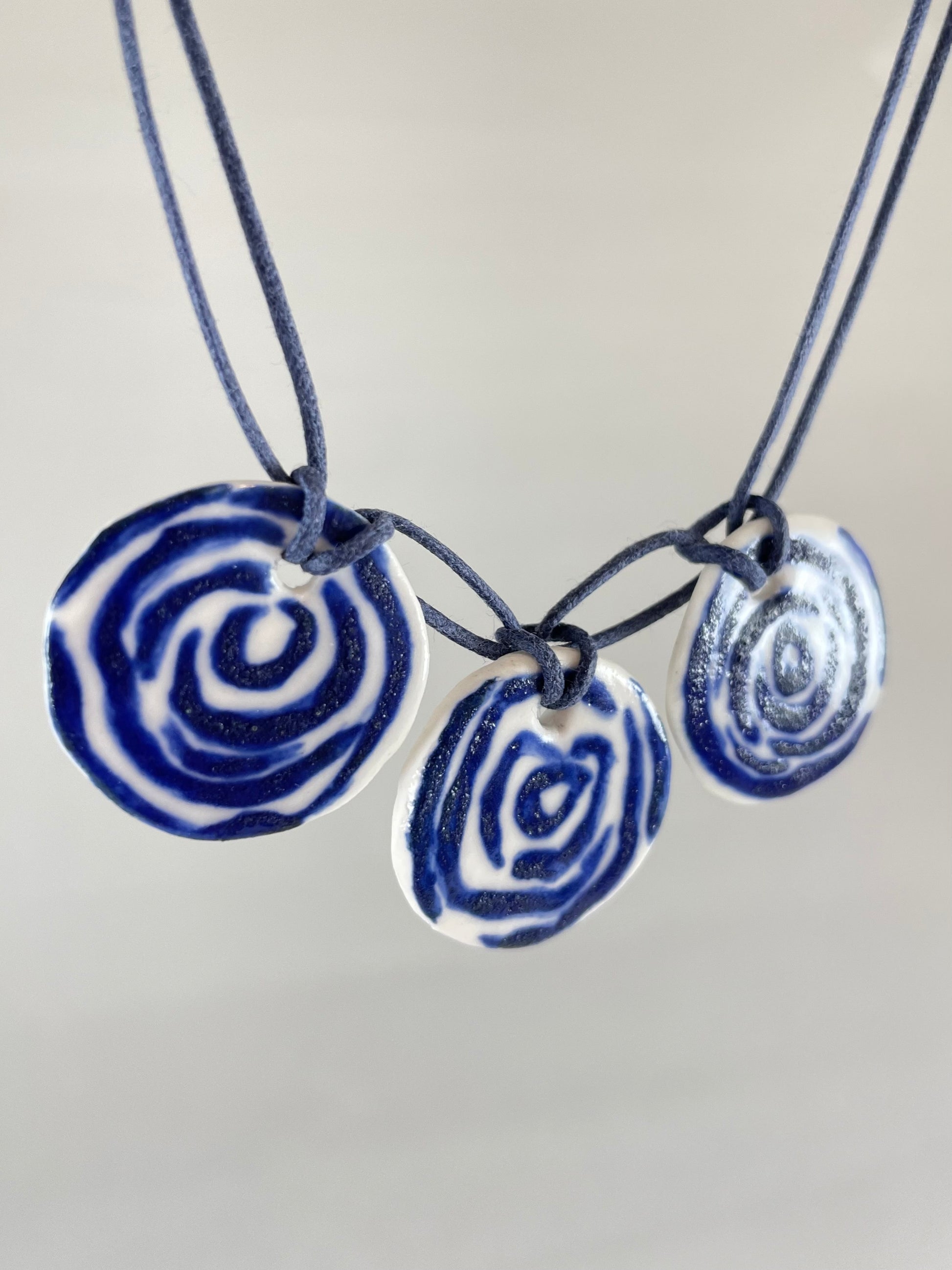 Necklace with three circular porcelain pendants on navy blue coloured thread, hanging in front of a white background. Blue rose pattern painted on white porcelain.  Photographed from left side to show thin profile of porcelain discs