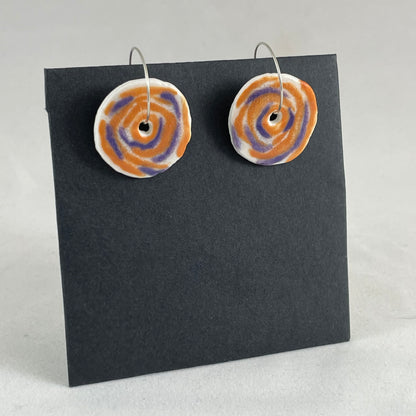A pair of lavender and orange porcelain earrings on a black background. Simple rose pattern. Side view.