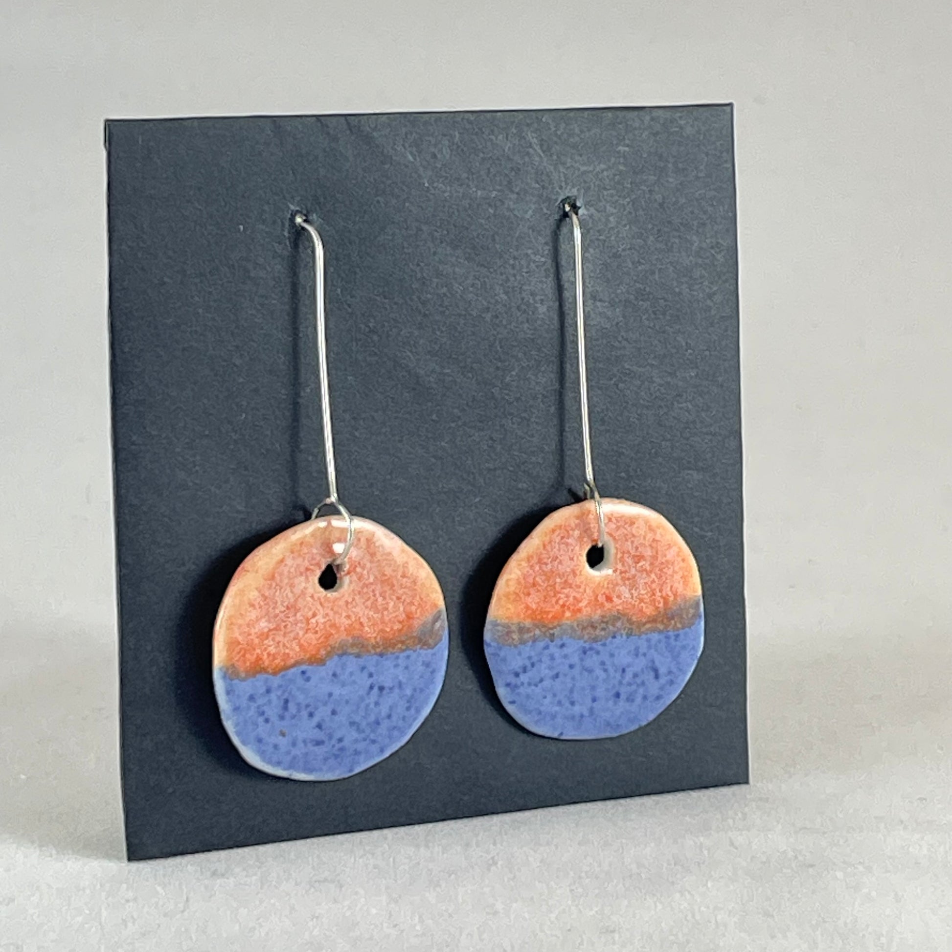 A pair of circular shaped porcelain earrings in blue and orange. On sterling silver earrings hooks. Side view