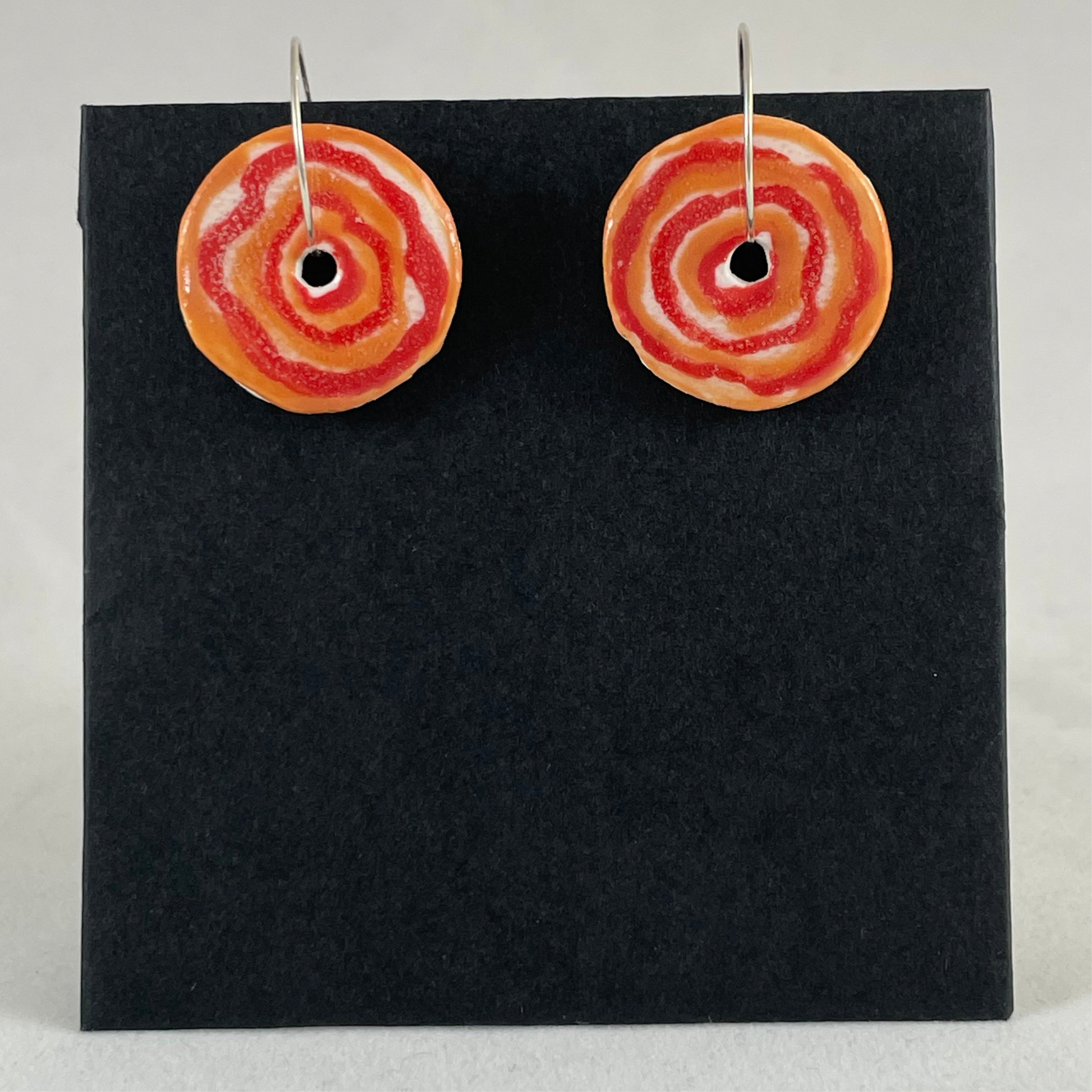 A pair of red and orange porcelain earrings on black card. box. Simple rose pattern with alternating, loosely painted red and orange circles.