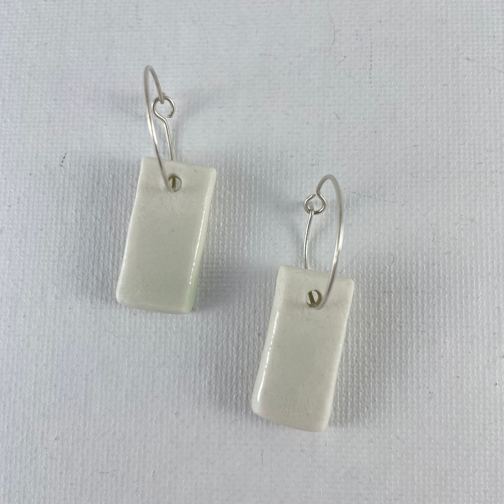 A pair of silver hoops with hand formed white porcelain pendants laid flat on a white background.