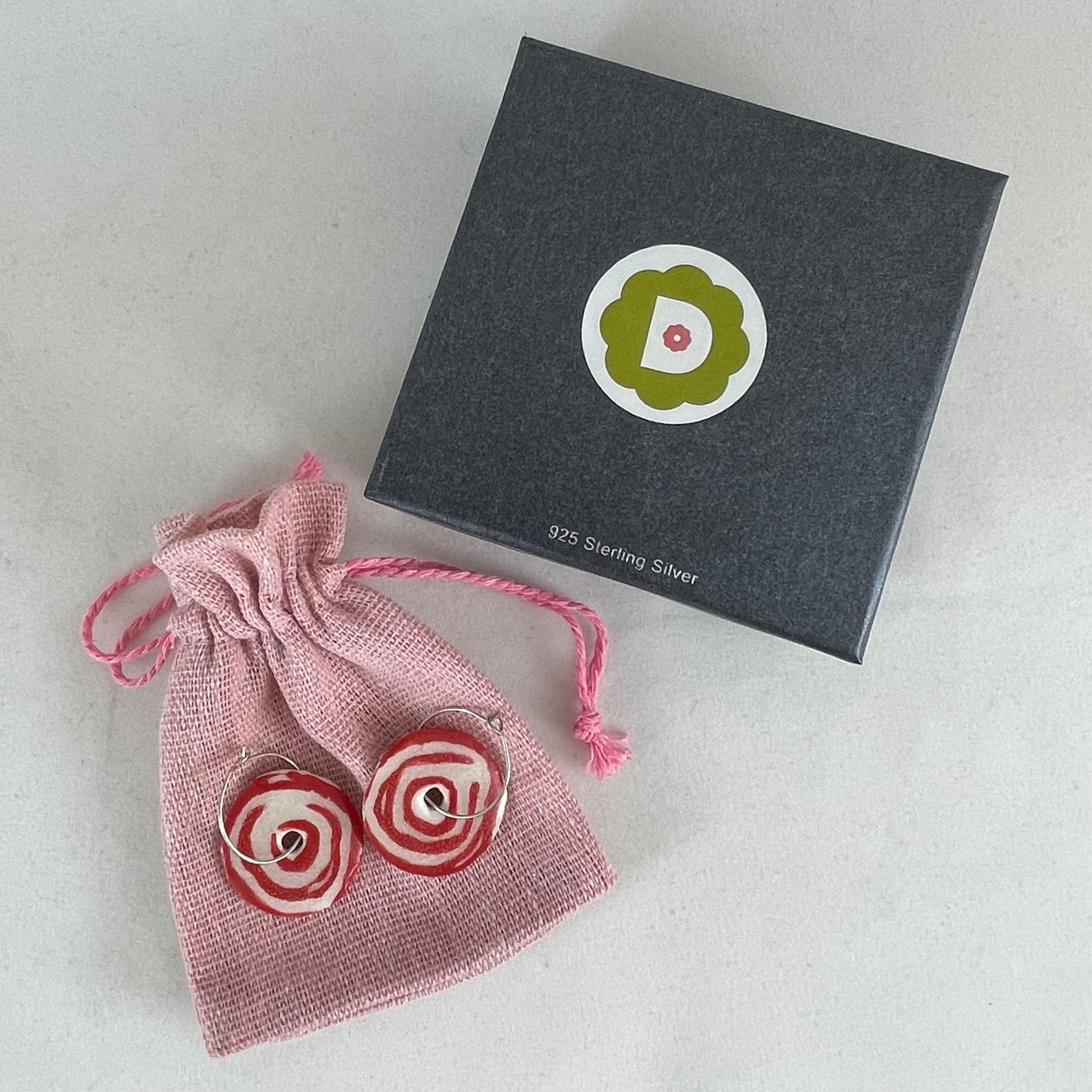 A pair of red and white porcelain earrings displayed next to dark grey jewellery box and pink linen pouch.