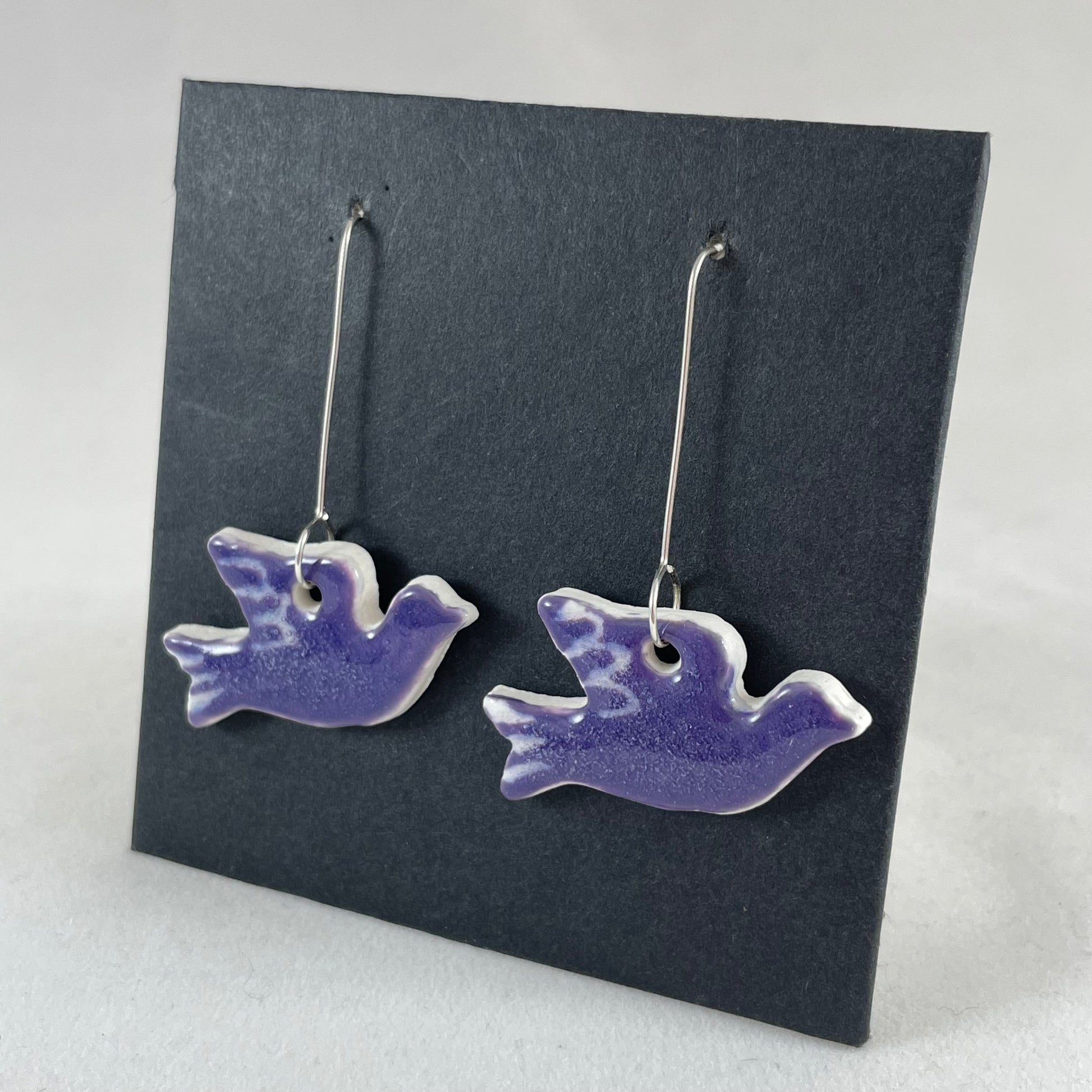A pair of lavender porcelain earrings in the shape of flying dove on a black background. Side view