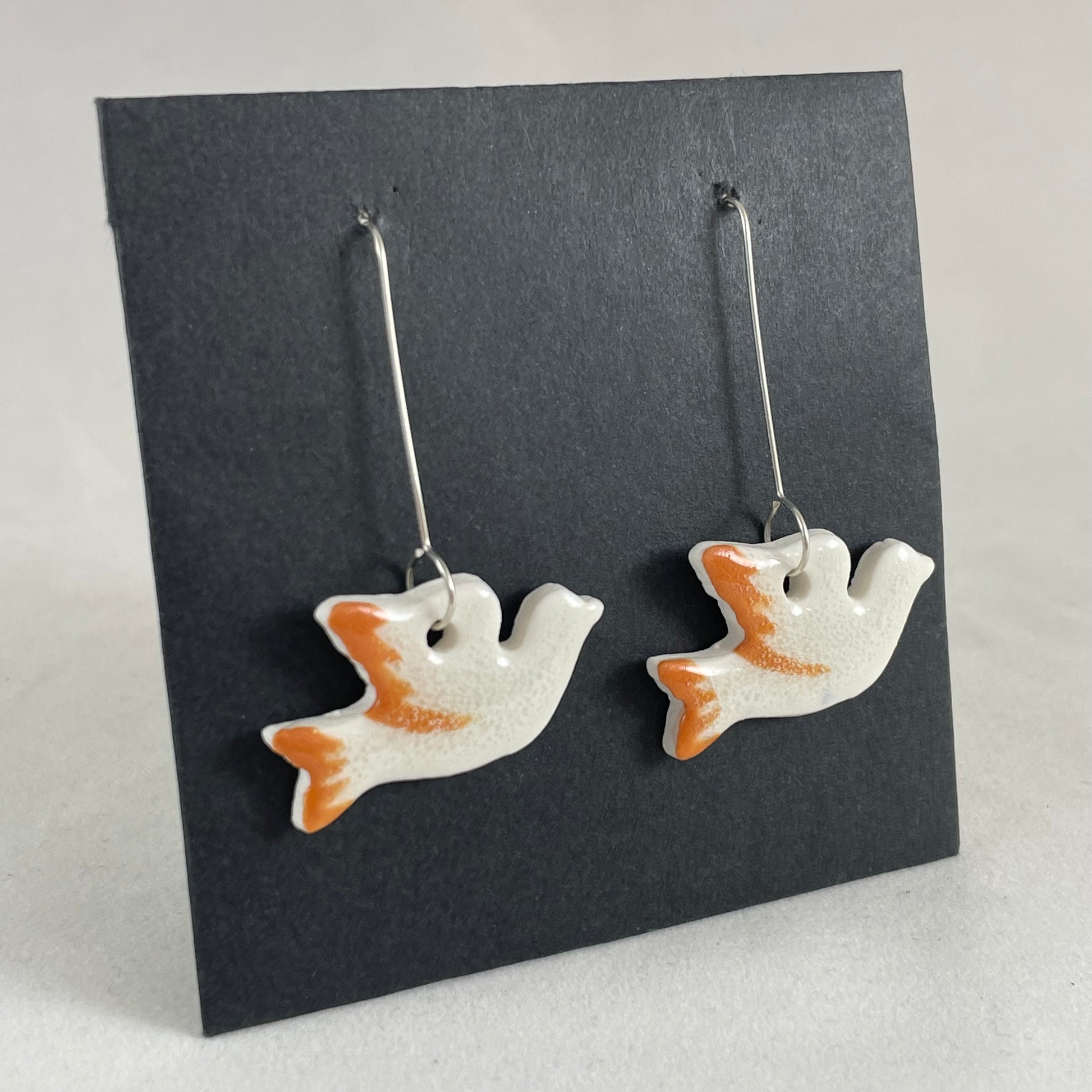 A pair of white and orange porcelain earrings in the shape of flying dove on a black background. Side view
