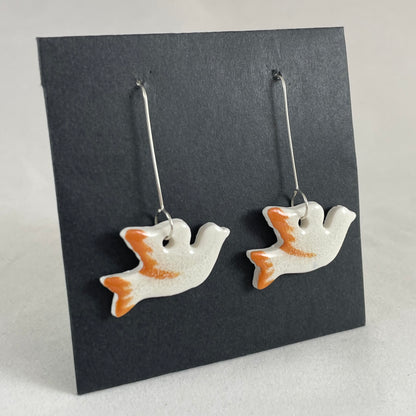 A pair of white and orange porcelain earrings in the shape of flying dove on a black background. Side view