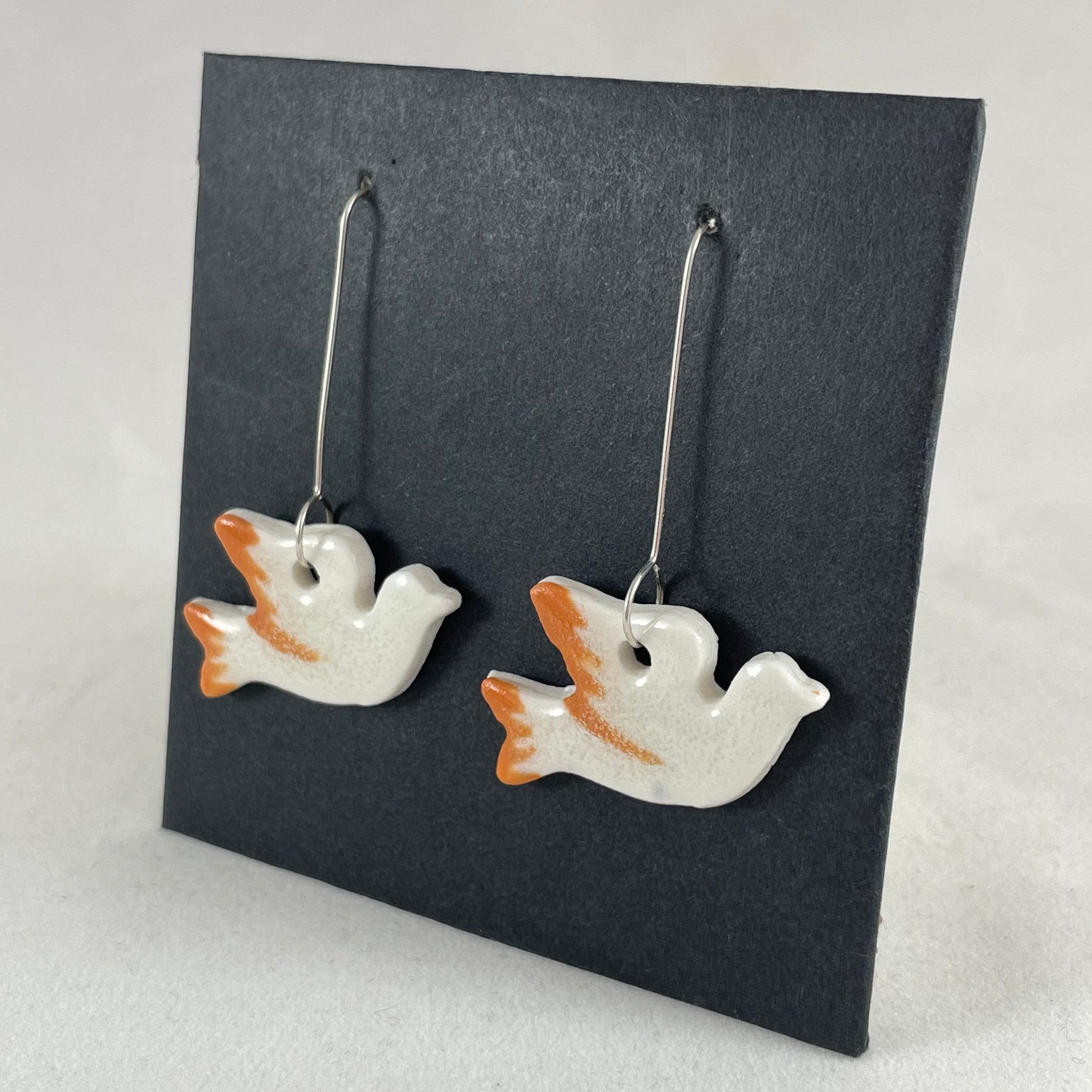 A pair of white and orange porcelain earrings in the shape of flying dove on a black background. Side view