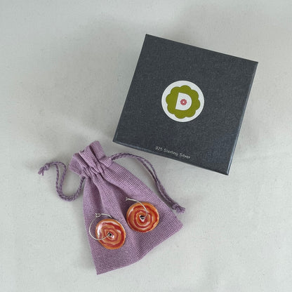 A pair of red and orange porcelain earrings displayed next to dark grey jewellery box. Earrings are resting on a lilac coloured linen jewellery pouch.