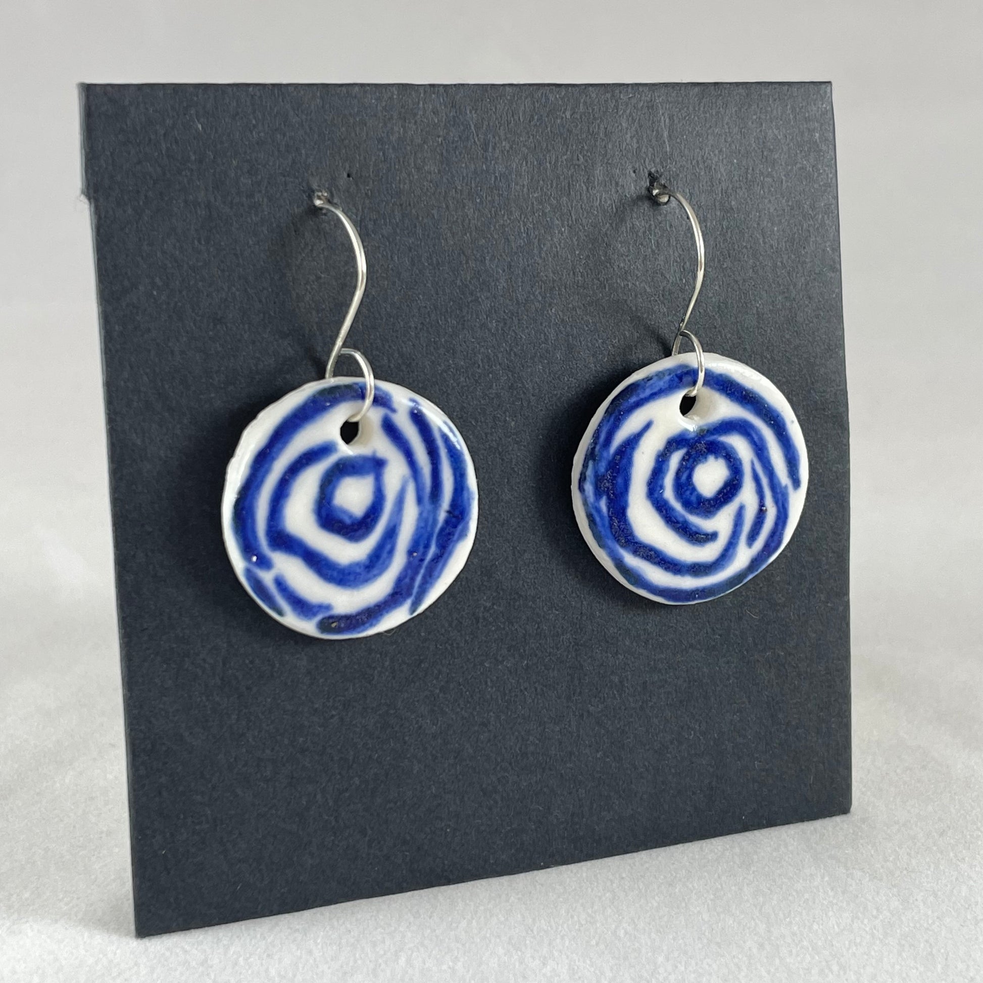 A pair of blue and white porcelain earrings hanging on black card. Simple rose pattern with loosely painted blue circles on white porcelain. Photographed to show earrings from left hand side 