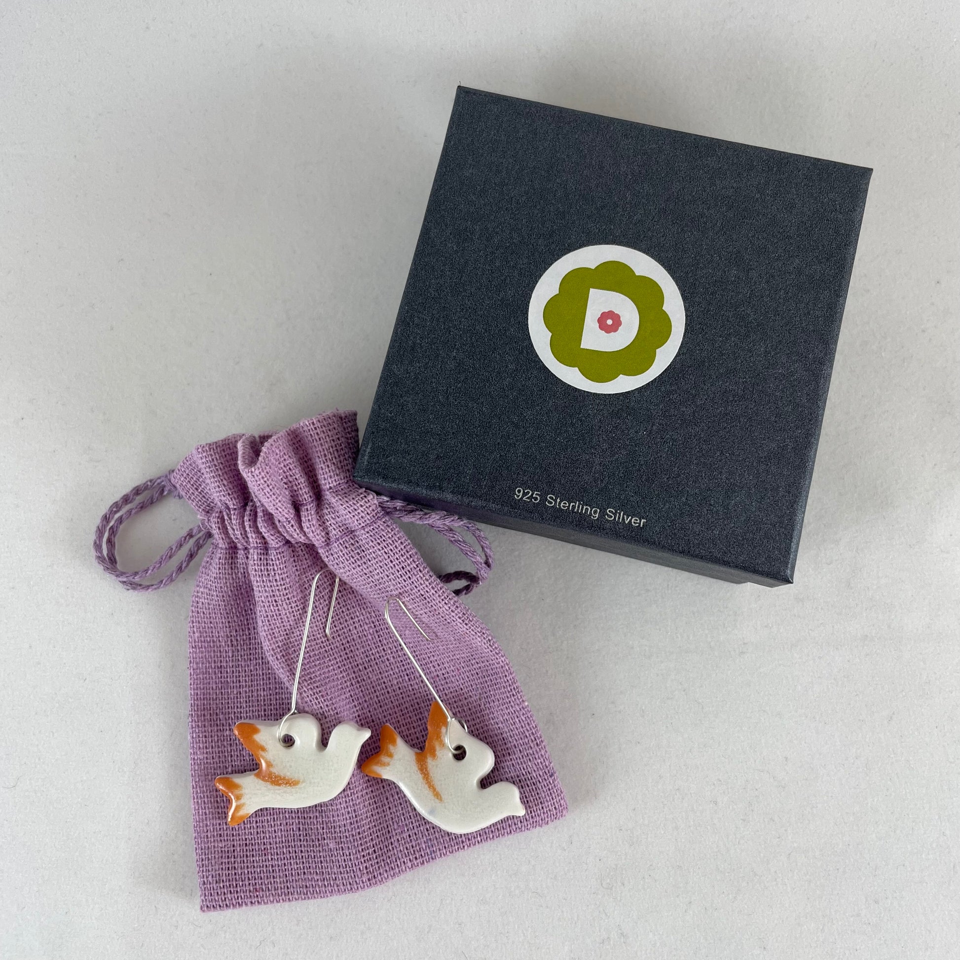 A pair of white and orange porcelain earrings in the shape of flying dove. Photographed on a linen pouch next to jewellery box.