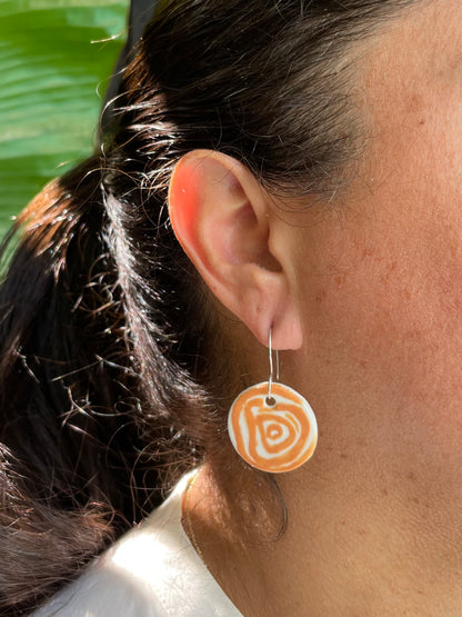 Closeup of woman with tan skin wearing white and orange circular earrings with rose pattern. Woman has her dark brown hair is pulled back in ponytail. A bright green bird of paradise plant is partially visible in the background   