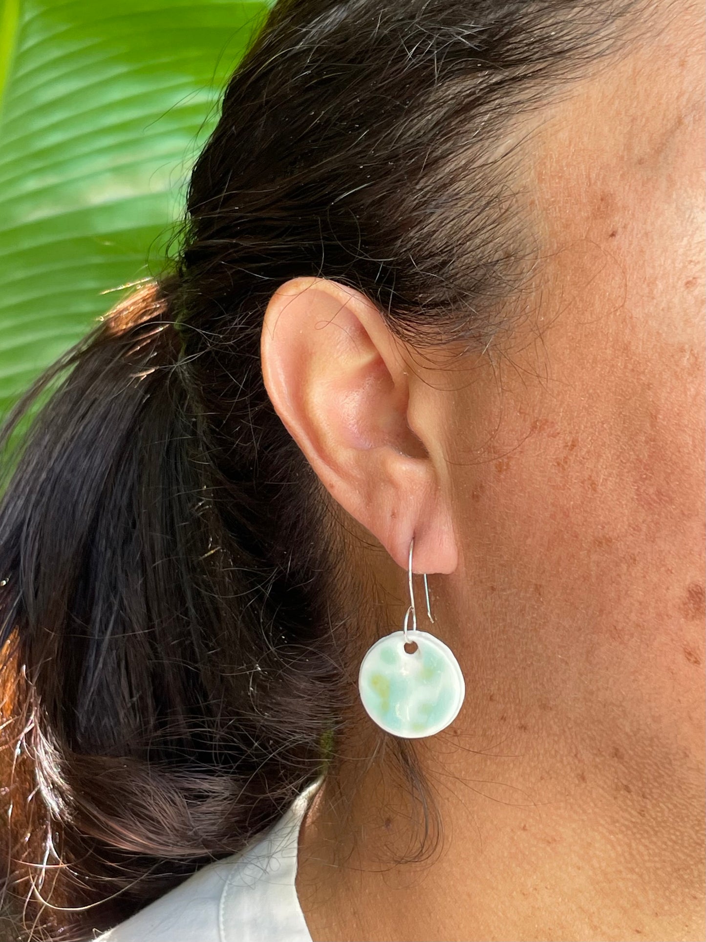 Closeup of woman with tan skin wearing mint, olive green and white and circular earrings. Woman has her dark brown hair is pulled back in ponytail.